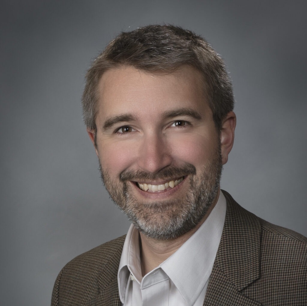 Tim's headshot. He is wearing a brown suit and white drass shirt, smiling in front of a grey background