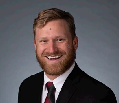 Charlie's headshot. He is in a black suit and dark red tie smiling in front of a grey background