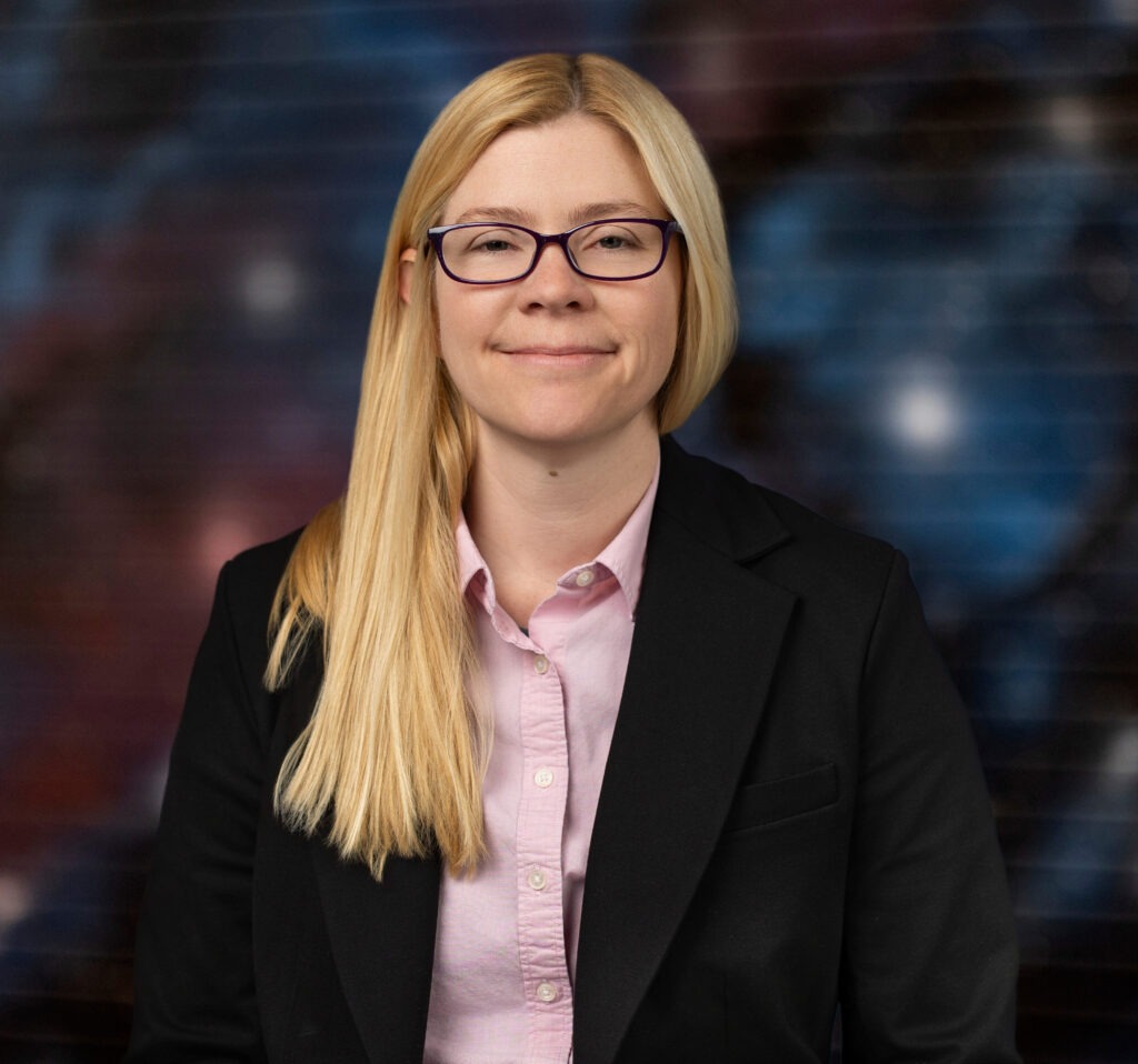 Sarah's headshot. She is wearing a black suit jacket with a light pink button down shirt. She is in front of a blurred image of what looks to be a galaxy